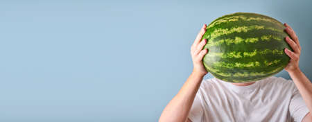 Young man keep whole watermelon in hands in white t-shirt on blue background. Mock up for design. Copy space.の写真素材