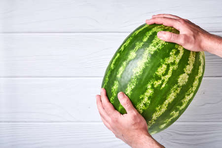 Young man keep whole watermelon in hands in white t-shirt on white wooden background. Mock up for design. Copy space.の写真素材