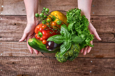 Female hand holding a basket with fresh vegetables and fruits on old wooden rustic background, top...