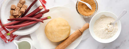 Ball of dough and ingredients flour, water, butter, sugar and rhubarb for baking on white marble background with dusting of flour. Recipe step by step. Selective focus. Top view. Banner.の写真素材
