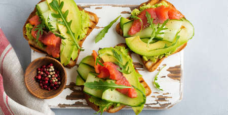Open toast with trout, salmon, avocado, cucumber and arugula on wooden stand on gray concrete...