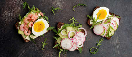 Open sandwiches on dark rye bread with eggs, shrimps, radishes, cucumber, cream cheese and arugula for breakfast on old dark background. Smorrebrod Traditional dish of Danish cuisine. Top view.の写真素材