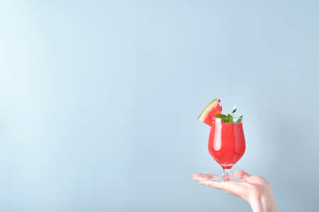 Female hand holds watermelon cocktail in glass with mint and ice on blue background. Colorful hot summer holidays. Close up slices of watermelon in hands. Banner. Mock up.の写真素材