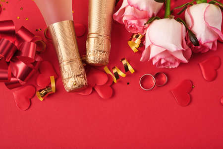 Pink roses flowers, wedding rings, champagne, gift, golden ribbons and confetti hearts on red...