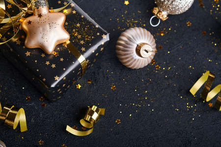 Holiday gift box or present with ribbon, golden confetti and gold baubles on black background. Magic...