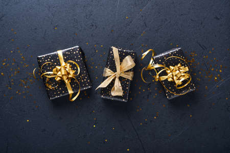 Holiday gift box or present with ribbon, golden confetti and gold baubles on black background. Magic christmas greeting card. Christmas Decoration. Border design. Mock up. Top view.の写真素材