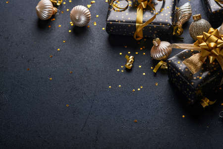 Holiday gift box or present with ribbon, golden confetti and gold baubles on black background. Magic christmas greeting card. Christmas Decoration. Border design. Mock up. Top view.の写真素材