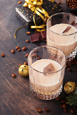 Glass of Irish cream barley liqueur with ice, roasted coffee beans, cinnamon, Christmas decoration and chocolate on dark wood background table. Christmas table with gifts. Selective focus.の写真素材