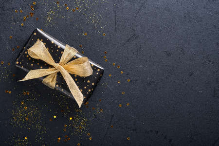 Holiday gift box or present with ribbon, golden confetti and gold baubles on black background. Magic christmas greeting card. Christmas Decoration. Border design. Mock up. Top view.の写真素材