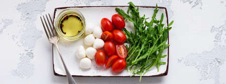 Mozzarella, cherry tomatoes and arugula served in white ceramic rectangular plates with olive oil over gray texture background. Flat layの写真素材
