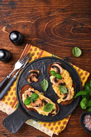 Scrambled eggs with fried mushrooms and basil on bread on black table background. Homemade breakfast or brunch meal - scrambled eggs and mushrooms sandwiches. Top view with copy spaceの写真素材