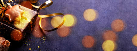 Holiday gift box or present with ribbon, golden confetti and gold baubles on black background. Magic christmas greeting card. Christmas Decoration. Toned image with bokeh. Mock up. Banner.の写真素材