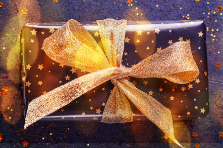 Holiday gift box or present with ribbon, golden confetti and gold baubles on black background. Magic christmas greeting card. Christmas Decoration. Toned image with bokeh. Mock up. Banner.の写真素材