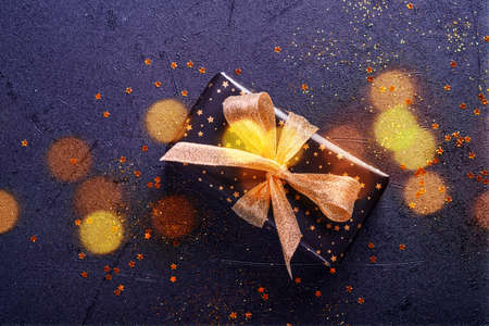 Holiday gift box or present with ribbon, golden confetti and gold baubles on black background. Magic...