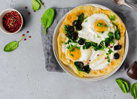 Turkish eggs flatbread with yoghurt, cheese, olives, spinach and red pepper on ceramic vintage plate on gray old background. Top view.の写真素材