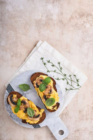 Scrambled eggs with fried mushrooms and basil on bread on white table background. Homemade breakfast...