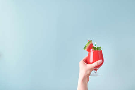 Female hand holds watermelon cocktail in glass with mint and ice on blue background. Colorful hot summer holidays. Close up slices of watermelon in hands. Banner. Mock up.の写真素材