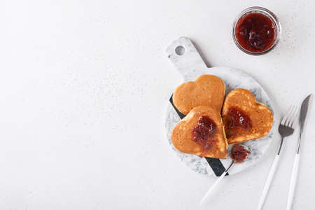 Pancakes with berry jam and honey in shape of heart and hot cup of coffee over white wooden background. Concept breakfast for Valentines Day or pleasant surprise for loved one Table viewed from aboveの写真素材