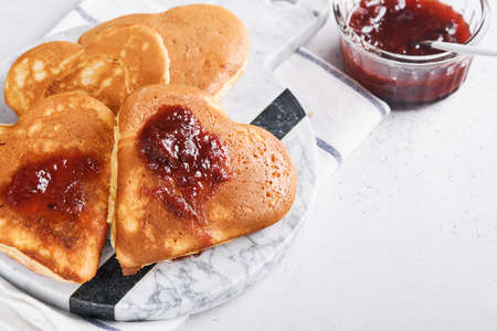 Pancakes with berry jam and honey in shape of heart and hot cup of coffee over white wooden background. Concept breakfast for Valentines Day or pleasant surprise for loved one Table viewed from aboveの写真素材