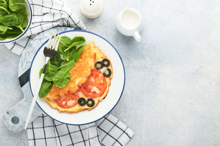 Omelette with tomatoes, black olives, cottage cheese and green spinach herbs on white plate on gray...