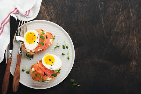 Sandwich with delicious toppings smoked salmon, eggs, herbs and microgreens radish, black sesame seeds over white plate on old wooden table background. Healthy open sandwich superfood. top view.の写真素材