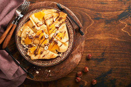 Crepes suzette with oranges and chocolate. Thin crepes with chocolate spread, hazelnuts and orange slices fruit in plate for breakfast on old dark wooden rustic table background. copyspace. top viewの写真素材