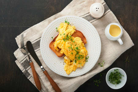 Scrambled Eggs. Fluffy and Buttery scrambled eggs on bread with microgreen radish and hollandaise sauce on white plate over dark old wooden background. Homemade breakfast or brunch meal. top view.の写真素材