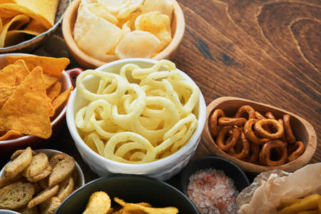Unhealthy foods or snacks. All classic potato snacks with peanuts, popcorn and onion rings and salted pretzels in bowl plates on old wooden background. Unhealthy food for figure, heart, skin, teeth.の写真素材