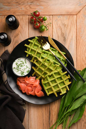 Green Belgian waffles. Spinach or wild garlic or pesto waffles with red salmon and cream sauce on black plate over dark rustic wooden background. Delicious breakfast, snack, brunch. top view. Mock upの写真素材