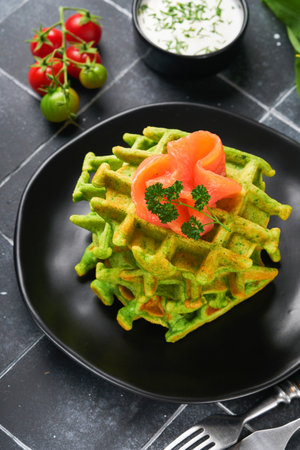 Green Belgian waffles. Spinach or wild garlic or pesto waffles with red salmon and cream sauce on old black tile table background. Delicious breakfast, snack, brunch on dark background.の写真素材