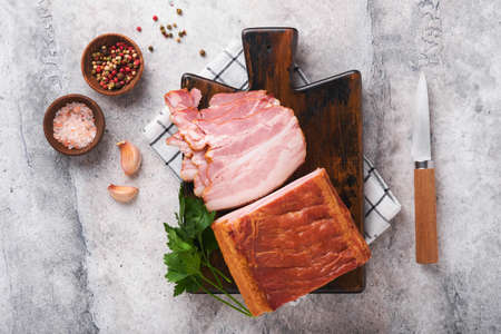 Whole Smoked Slab Bacon. Brisket. Sliced smoked gammon on a wooden table with rosemary, parsley, pepper, salt, and garlic on old gray concrete table background. copyspace.の写真素材
