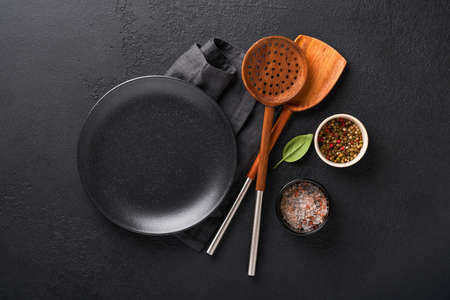 Cooking wooden utensils, black empty plate, basil leaves and spices on dark stone background....