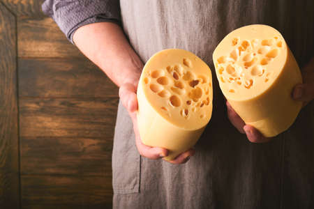 Hands of man hold two big slices of cheese maasdam against background of old wooden wall in cheese factory. Cheesemaker hold cheese maasdam in hand. Free space for your text.の写真素材