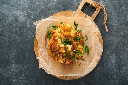 Baked cauliflower. Oven or whole baked cauliflower spices and herbs server on wooden rustic board on old gray background table. Delicious cauliflower. Eyal Shani dish. Perfect tasty snack.の写真素材