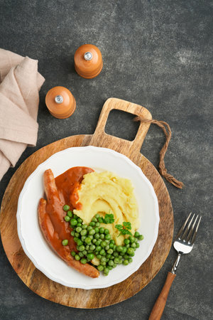 Bangers and mash. Grilled sausages with mash potato and green pea on white plate on black dark  background. Traditional dish of Great Britain and Ireland. BBQ beef sausages. Top view.の写真素材