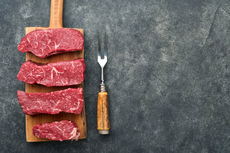 Raw steaks. Top blade steaks on wood burning board with spices, rosemary, vegetables and ingredients for cooking on black background. Top view. Copy space.の写真素材