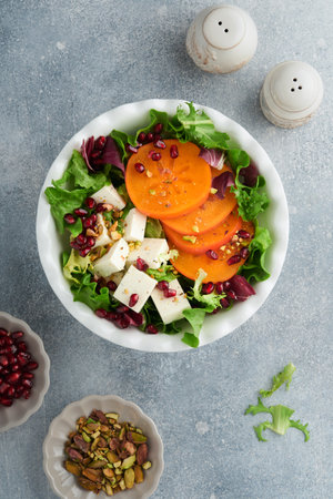 Salad. Fresh green salad with feta cheese, persimmon, pomegranate and pistachio on white plate. Idea for healthy delicious winter Christmas salad. Healthy balanced eating. top view.の写真素材