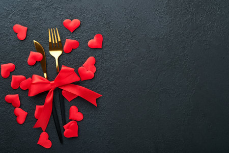 Valentines day romantic table setting. Empty or closeup of a dinner black plate, knife, fork and decorative silk hearts on black background. holiday concept. Copy space for inscriptions.の写真素材