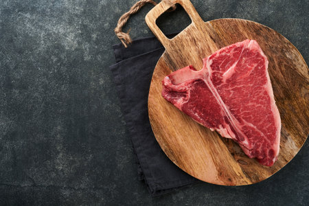 Thick Raw T-Bone Steak. Dry-aged Raw T-bone or porterhouse beef meat Steak on cutting boar with herbs and salt on dark background. Top view and copy space.の写真素材