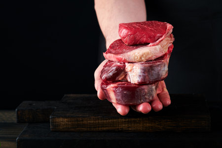 Chef cutting stack of beef meat from various or different steaks. Mans hands hold stack raw steak on rustic wooden cutting board on black background. Cooking, recipes and eating concept. Mock up.の写真素材