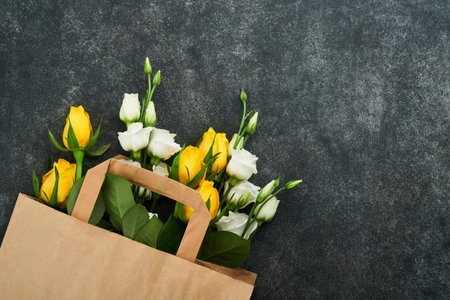 Easter concept. Bouquet of fresh white and yellow roses in paper bag on old dark concrete background. Passover Pesah celebration concept. Concept of mothers, womens day, spring background. top view.の写真素材