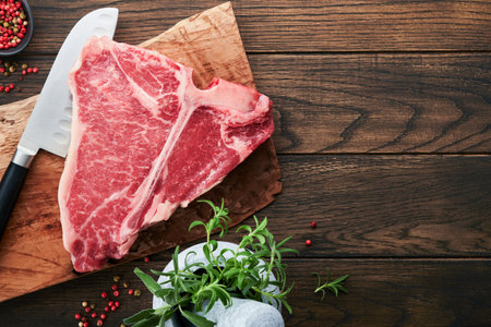 Thick Raw T-Bone Steak. Dry-aged Raw T-bone or porterhouse beef meat Steak on cutting boar with herbs and salt on dark background. Top view and copy space.の写真素材
