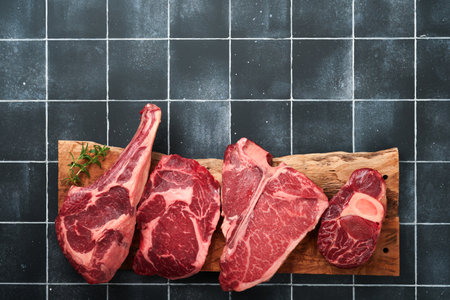 Raw prime steaks. Variety of fresh black angus prime meat steaks T-bone, New York, Ribeye, Striploin, Tomahawk on black old tiles stone background. Set of various classic steaks. Top view copy space.の写真素材