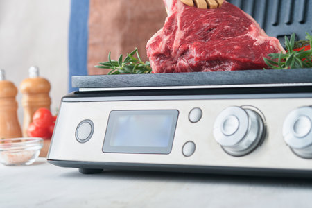 Raw prime steaks on electric grill. Chef male hand holding steak Ribeye on modern electric grill with rosemary, cherry tomato and pepper. Cooking process in modern kitchen. Top view, copy space.の写真素材