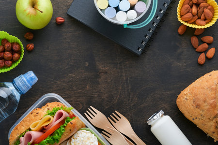 Healthy food for school lunch concept. Healthy bread, meat sandwich with cheese, apple, fresh cucumber, carrots, nuts in container, water and yogurt on dark background. Back to school concept.の写真素材