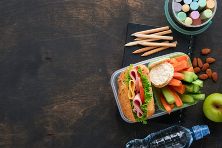 Healthy food for school lunch concept. Healthy bread, meat sandwich with cheese, apple, fresh cucumber, carrots, nuts in container, water and yogurt on dark background. Back to school concept.の写真素材
