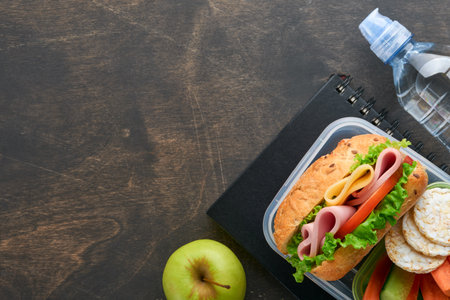 Healthy food for school lunch concept. Healthy bread, meat sandwich with cheese, apple, fresh cucumber, carrots, nuts in container, water and yogurt on dark background. Back to school concept.の写真素材