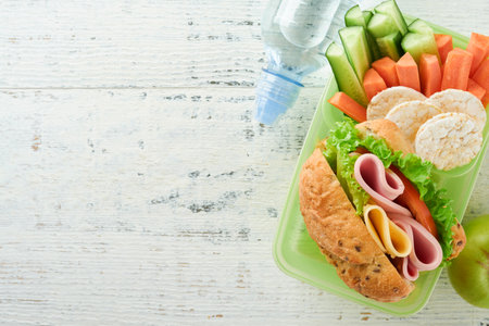 Healthy food for school lunch concept. Healthy bread, meat sandwich with cheese, apple, fresh cucumber, carrots, nuts in container, water and yogurt on kitchen background. Back to school concept.の写真素材