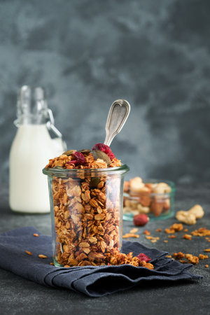 Homemade granola in glass jar with greek yogurt or milk and cashews, almonds, pumpkin with dried cranberry seeds in dark gray table background. Healthy energy breakfast or snack. top viewの写真素材