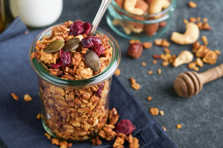 Homemade granola in glass jar with greek yogurt or milk and cashews, almonds, pumpkin with dried cranberry seeds in dark gray table background. Healthy energy breakfast or snack. top viewの写真素材